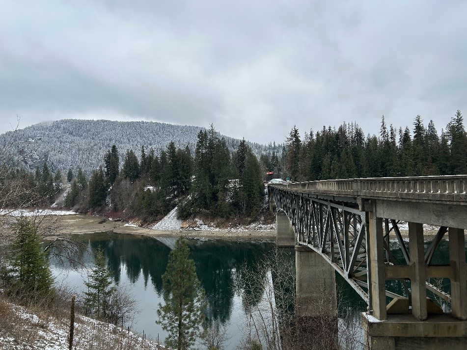 River with trees reflecting and a bridge. Snowy hills in the distance